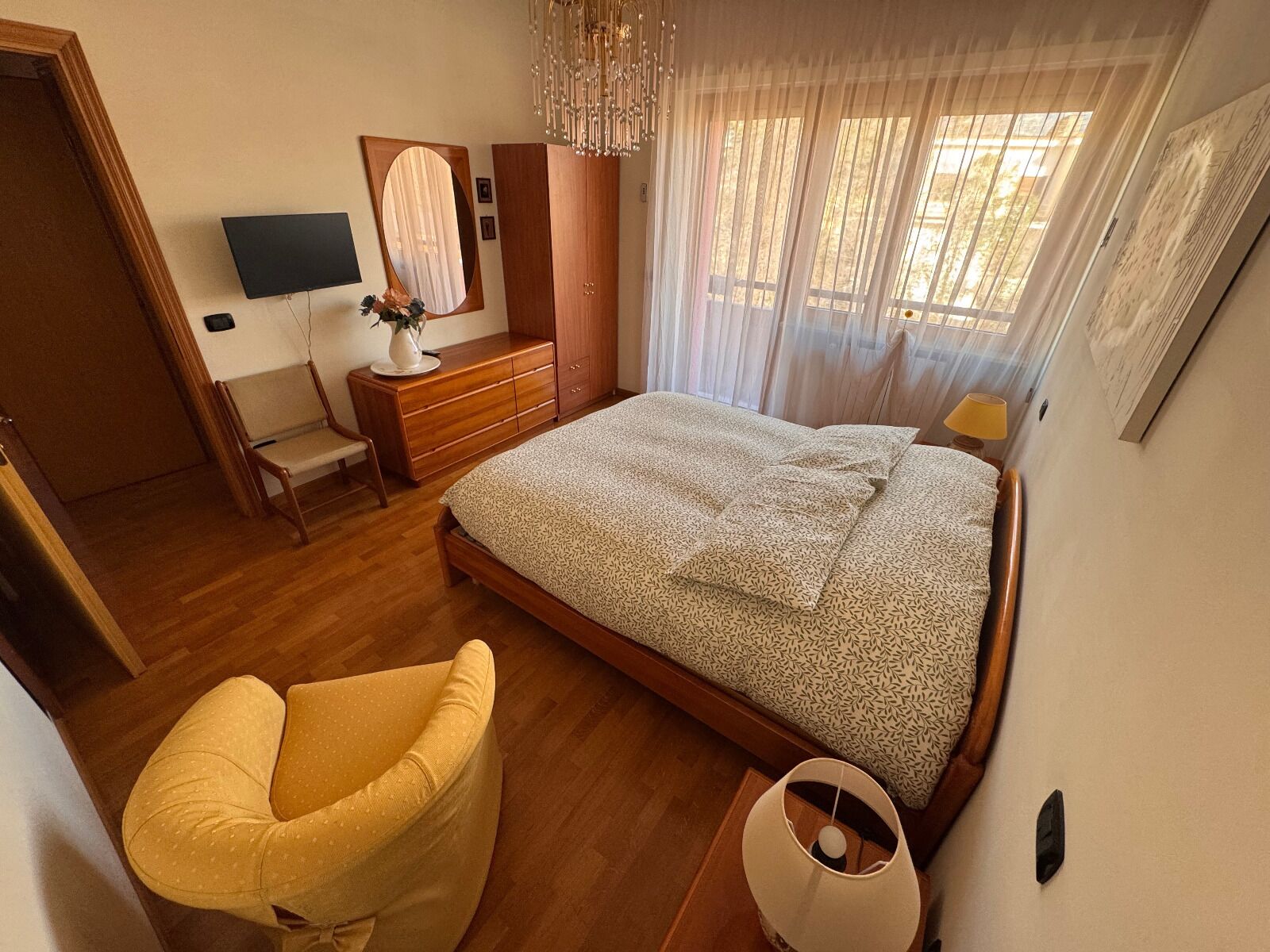 Un dormitorio con cama, una silla, cómoda, espejo, televisor y ventana con cortinas.