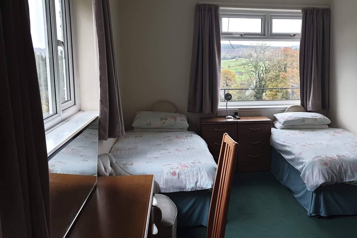 A small, tidy room with two single beds, a desk, and a chair. There is a window with curtains, a bedside table, and a trash bin.