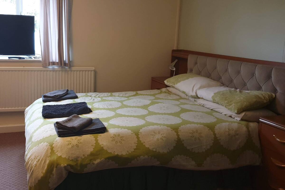 A hotel room with a bed, a television, a radiator, a bedside table, and a window.