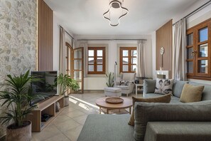 Ein helles Wohnzimmer mit Fernseher, Sofa und Couchtisch. Es befinden sich Pflanzen, eine Heizung und ein Fenster mit Vorhängen im Raum.
