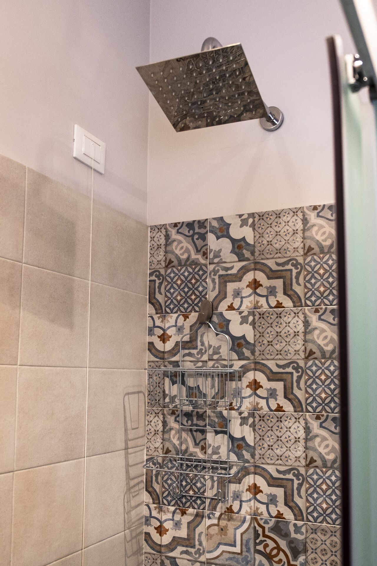 A shower with a square showerhead and a metal rack. The walls are tiled with patterned tiles.