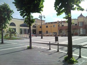 Une place agrémentée d’arbres, d’une fontaine et d’immeubles aux fenêtres en arc de cercle.