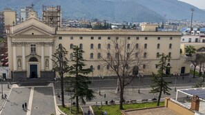 Un grand bâtiment à la façade classique, doté d’une entrée centrale et entouré d’une vaste place publique agrémentée d’arbres et de bancs.