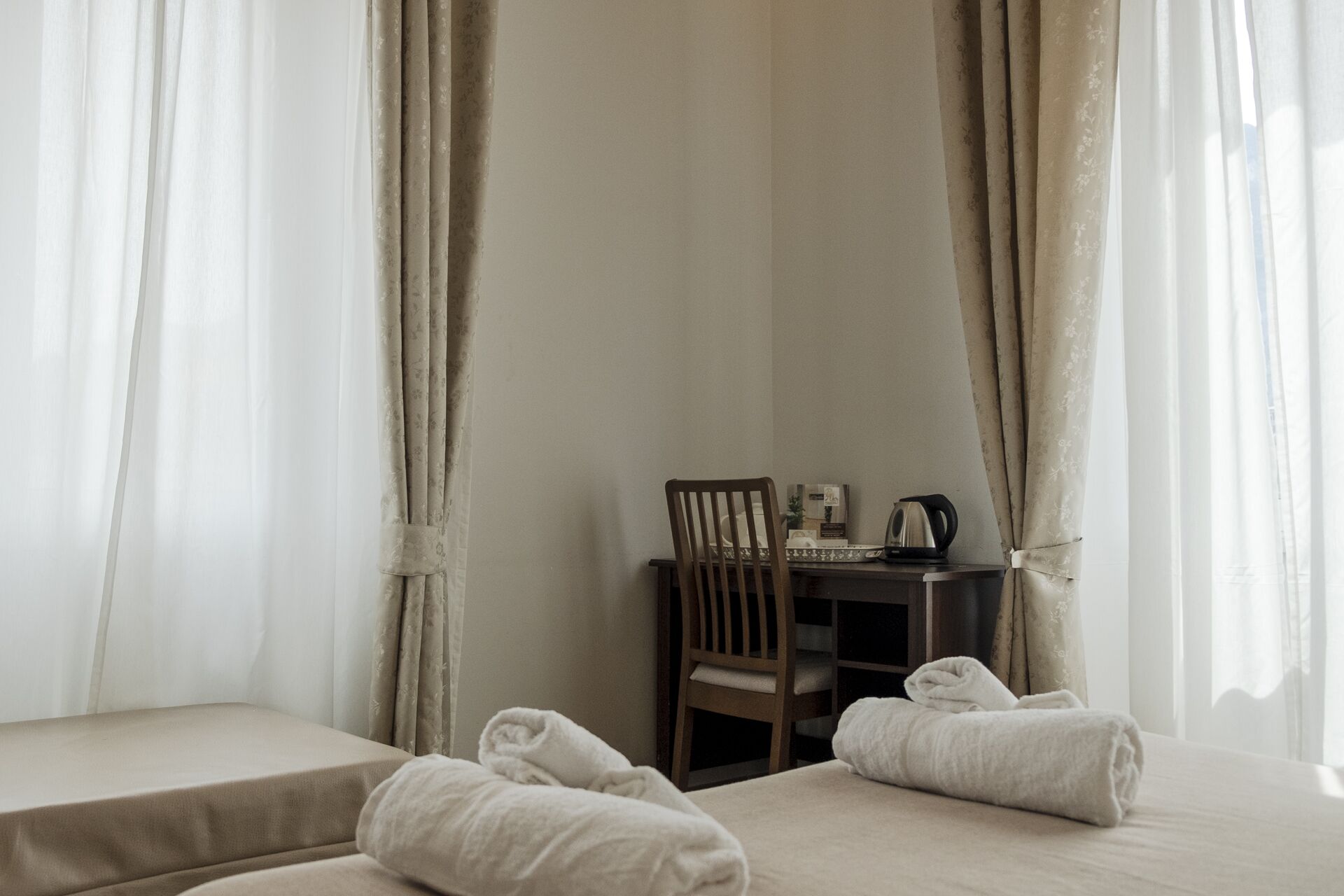A hotel room with two beds, a wooden chair, a small table with a kettle, and a framed picture.