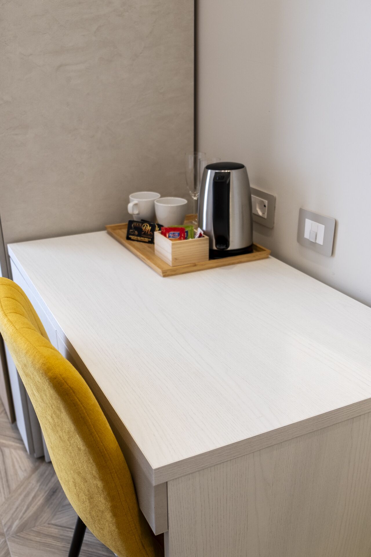 Un espacio de trabajo pequeño y ordenado con un escritorio de color claro, una silla amarilla, una cafetera, dos tazas y una caja de pañuelos.