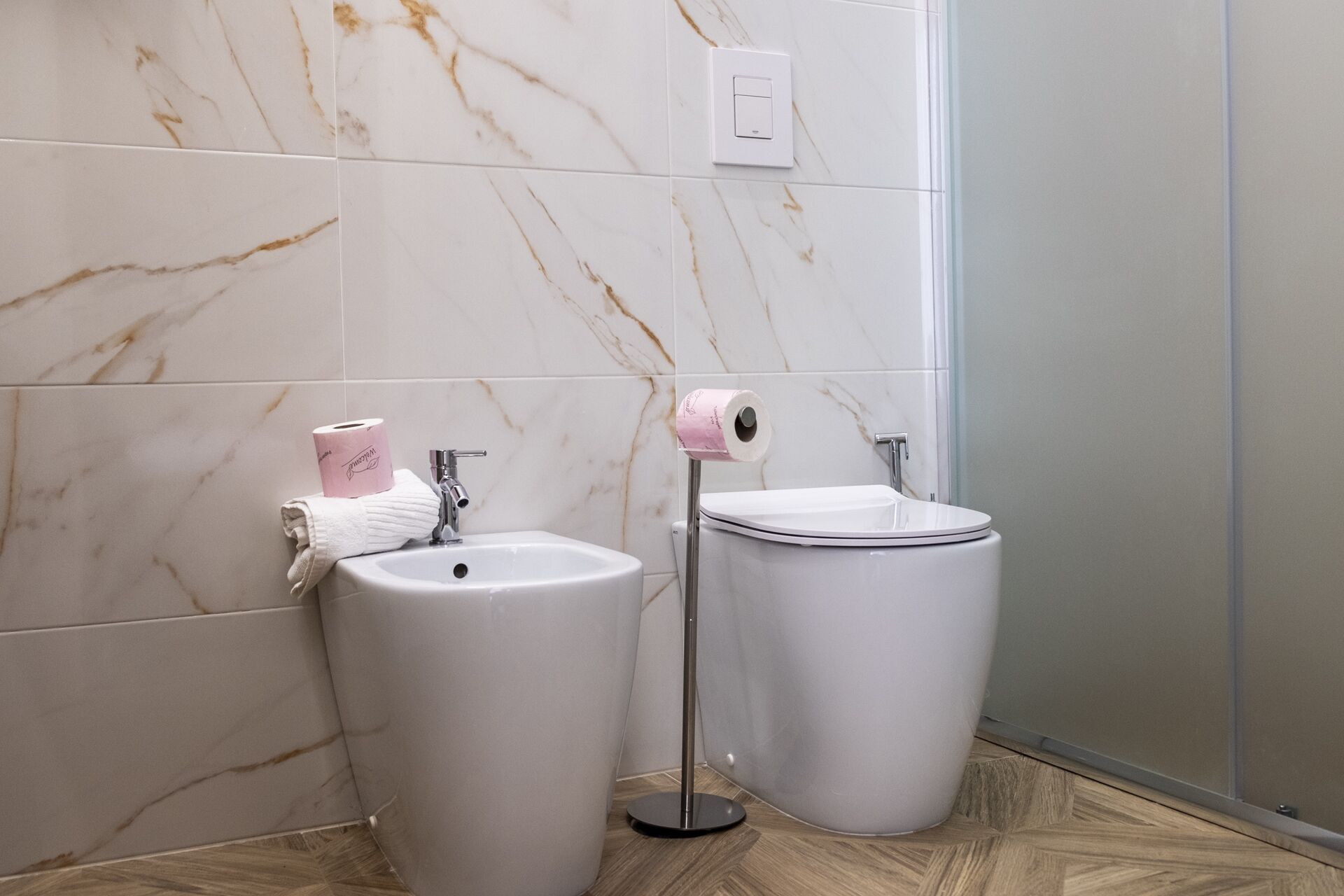 A bathroom with a marble wall, a sink, a toilet, and a roll of toilet paper on a holder.
