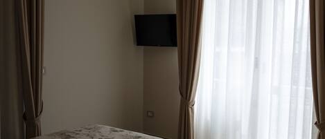 A hotel room with a bed, a flat-screen TV mounted on the wall, and sheer curtains on the window.