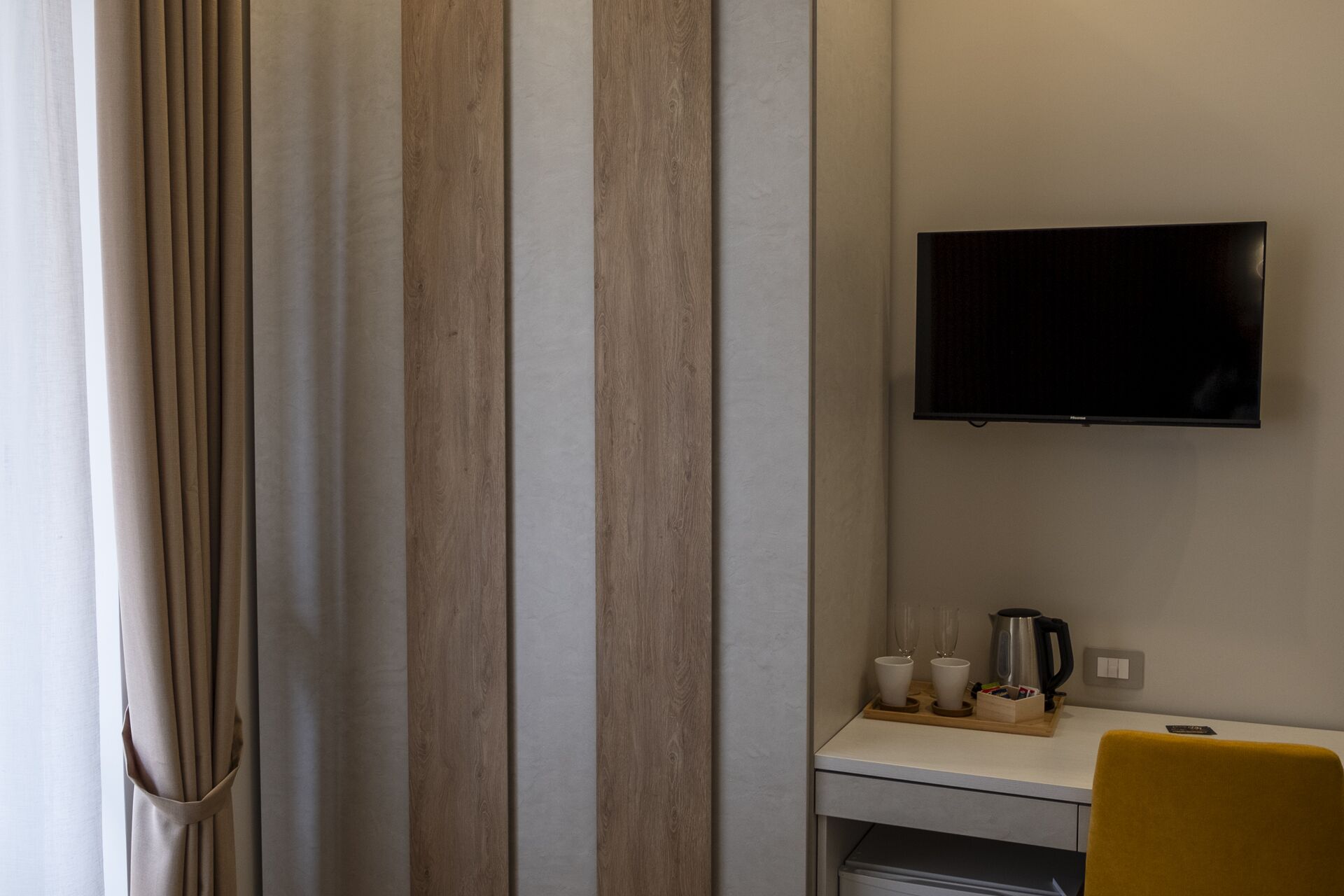 A hotel room with a TV mounted on the wall, a small table with cups and a kettle, and a yellow chair.