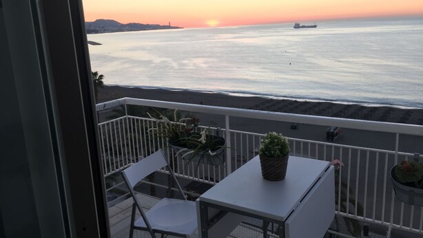 A balcony with a table and chairs, overlooking a beach with a sunset and a ship in the distance. - The Malagueta's Views (Málaga)