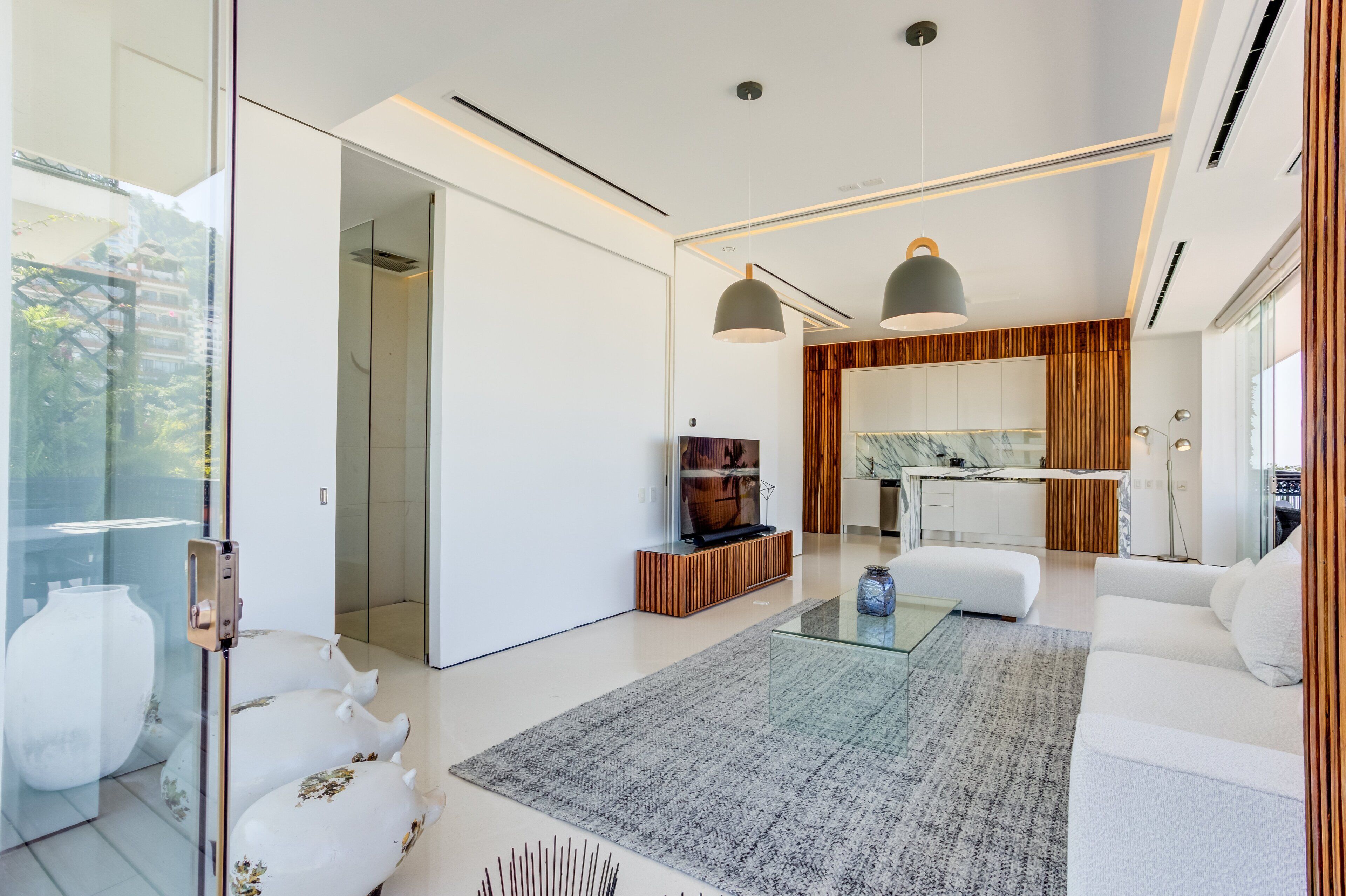 A modern living room with a glass coffee table, white sofas, and a kitchen area with wooden accents.