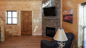 A cozy cabin interior with a stone fireplace, wooden walls, a television mounted on the wall, and a comfortable seating area.