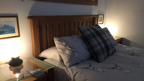 Un lit bien fait, recouvert d’une couvre-lit à motifs floraux, avec une tête de lit en bois et une table de chevet sur laquelle se trouvent une lampe et un livre.