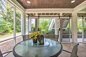 A round glass table with a flower vase and a lantern, surrounded by chairs, in a covered outdoor area with large windows and a view of greenery. - Waveland Family House, 4-Minute Drive to the Beach (Waveland)