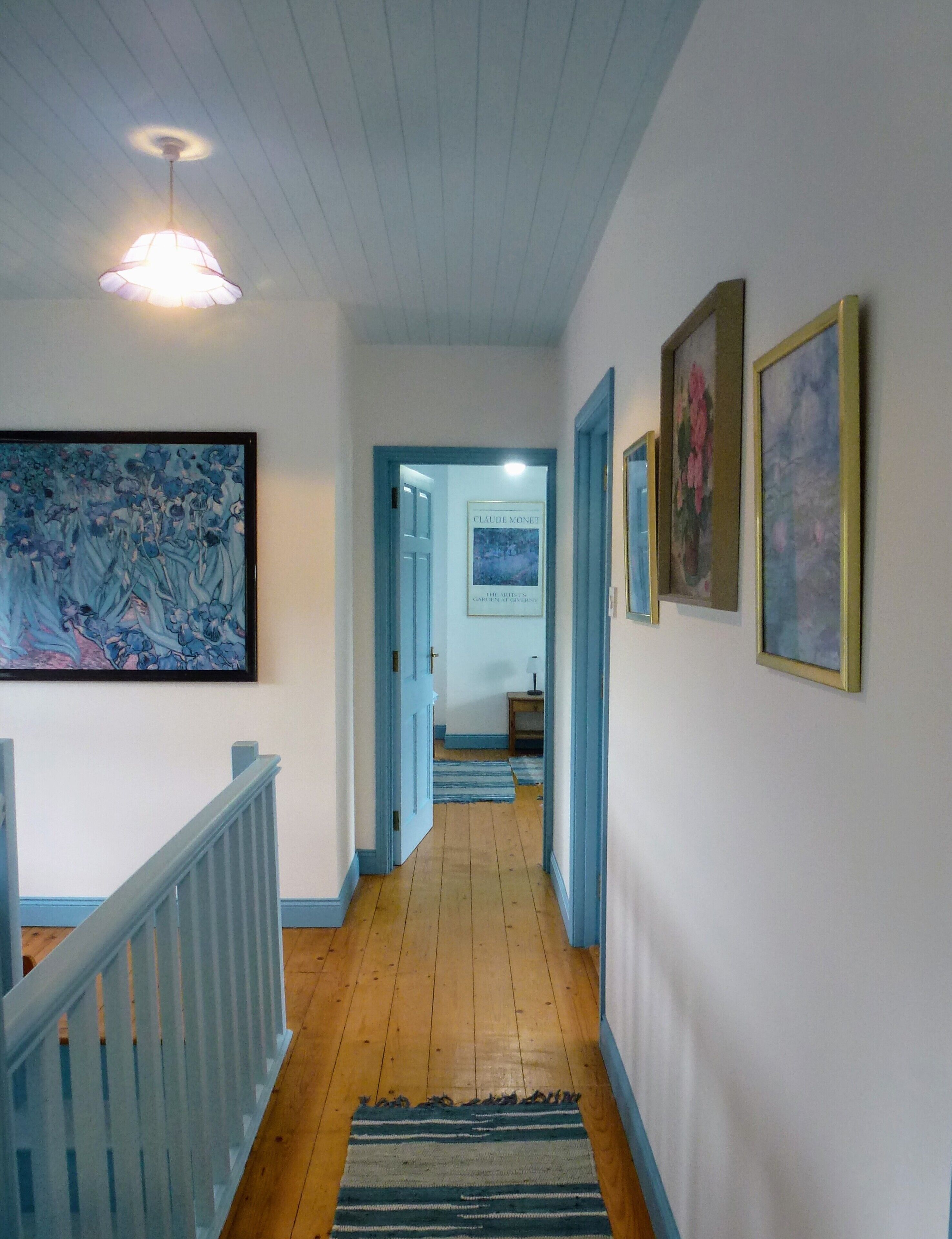 Un couloir doté d’un plancher en bois, de murs bleus et d’un escalier avec une rampe bleue. Des tableaux encadrés ornent les murs et un luminaire est fixé au plafond.