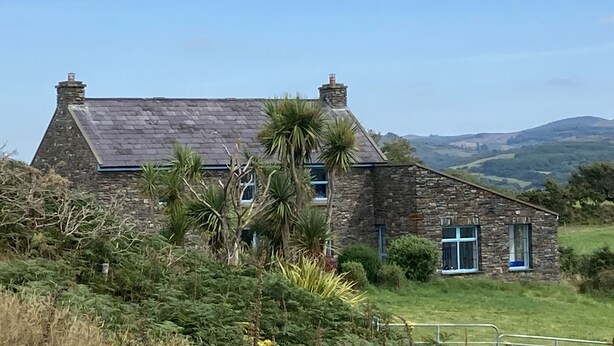 A stone house with a sloped roof and chimneys, surrounded by greenery and hills in the background.