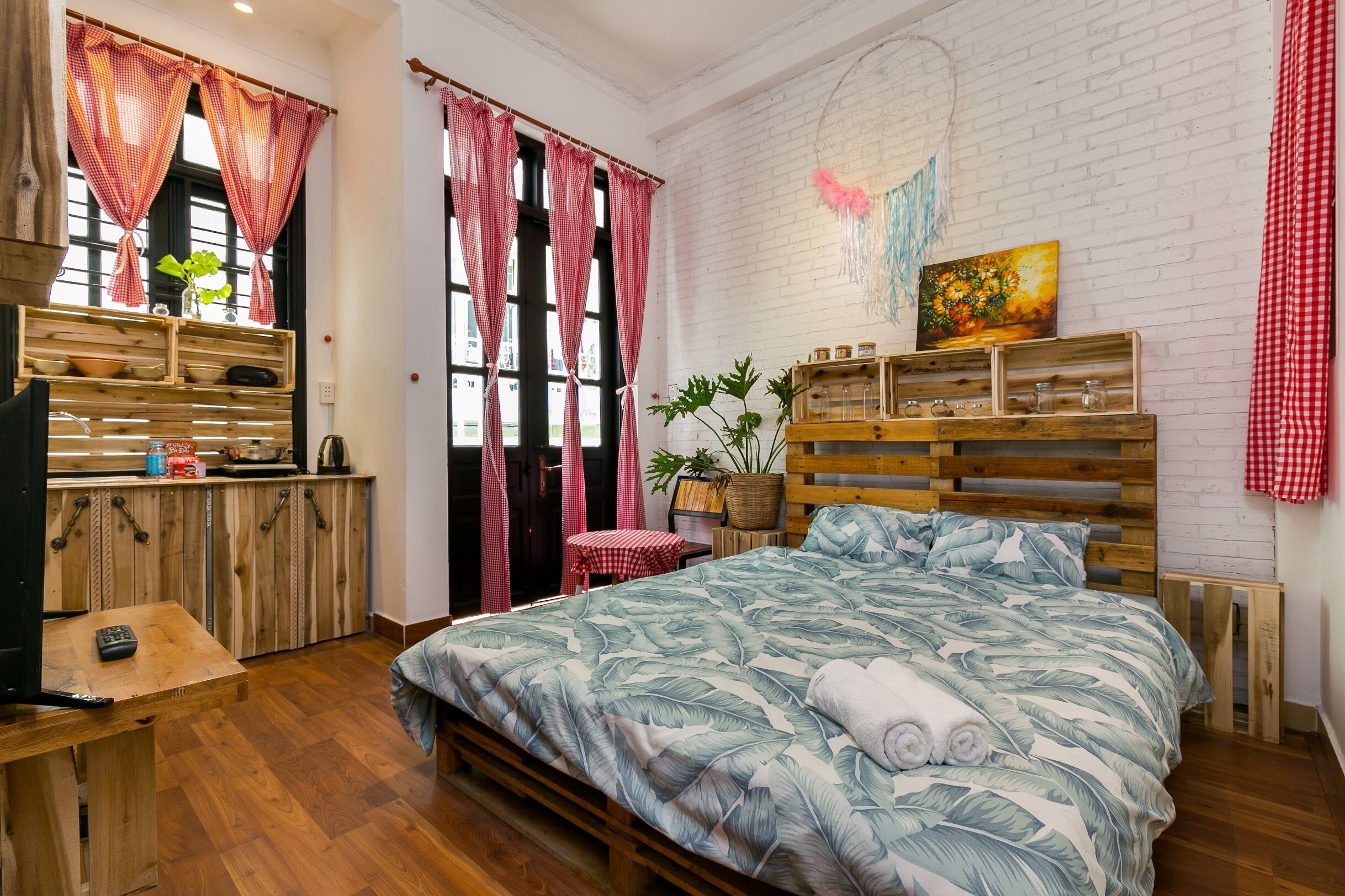 A cozy bedroom with a wooden bed, a small table, a red checkered tablecloth, and a brick wall with a painting.