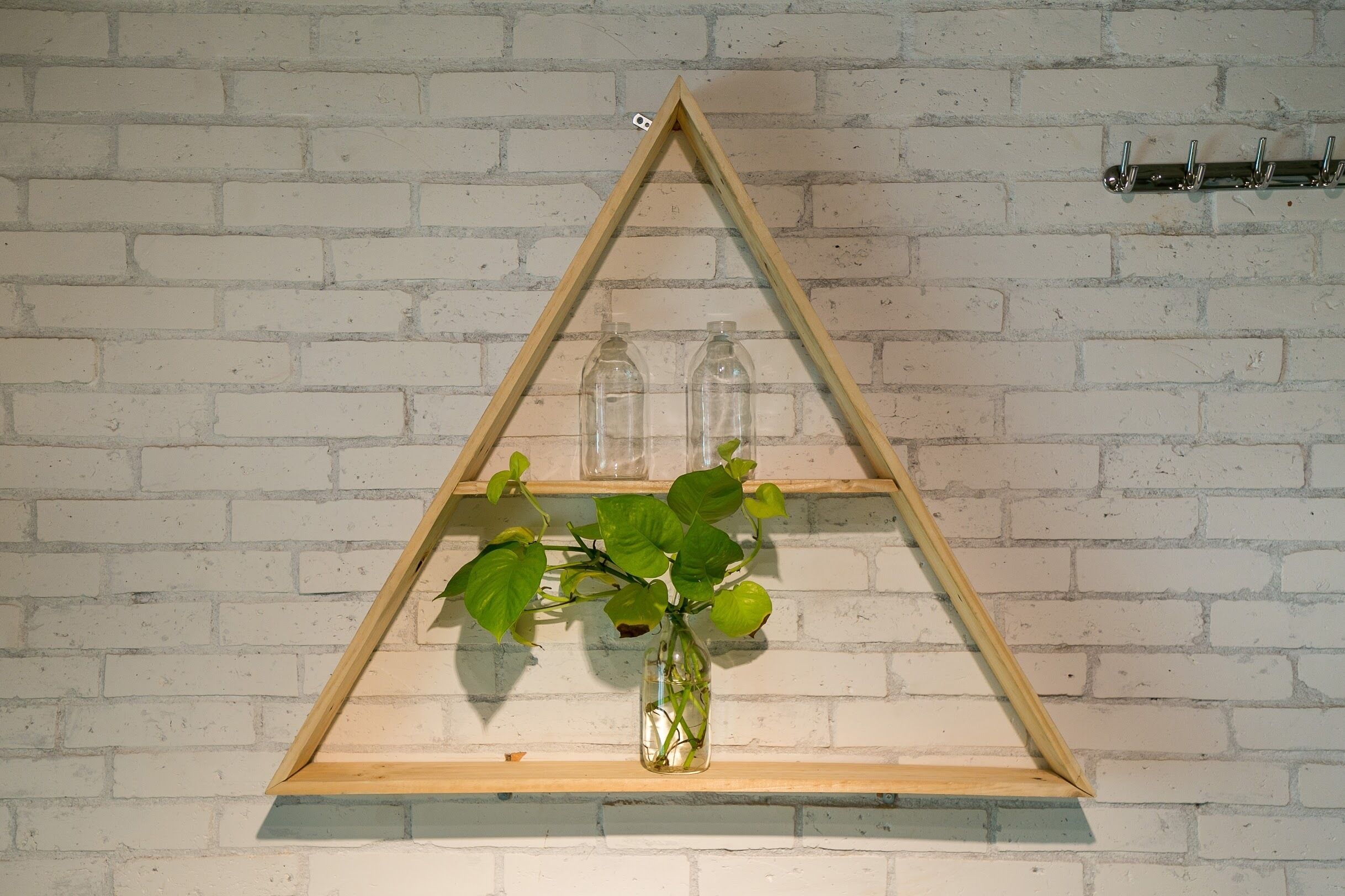 En triangulär trähylla mot en vit tegelvägg, med en vas med gröna blad och två tomma glasflaskor på.