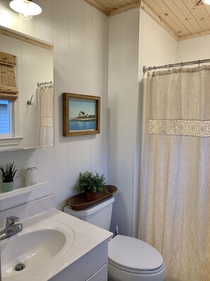 A bathroom with a white sink, toilet, and shower curtain. There is a framed painting on the wall and a potted plant on the counter. - Sunrise to Sunset - Steps From the Ocean! Hot Tub! (Kitty Hawk)