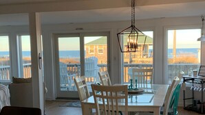Ein geräumiger Essbereich mit großem Tisch und Stühlen, große Fenster mit Blick auf Strand und Meer sowie eine Hängeleuchte über dem Tisch.