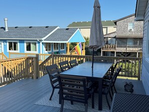 A deck with a table and chairs, a flag, and a building with a blue facade. - Sunrise to Sunset - Steps From the Ocean! Hot Tub! (Kitty Hawk)
