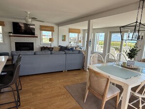 A modern living room with a grey sectional sofa, a dining table with chairs, a fireplace, and a large TV. - Sunrise to Sunset - Steps From the Ocean! Hot Tub! (Kitty Hawk)