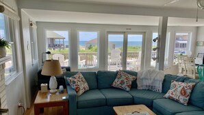 Ein Wohnzimmer mit einem türkisfarbenen Sofa, gemusterten Kissen, einem Holztisch und Blick auf den Strand durch große Fenster.