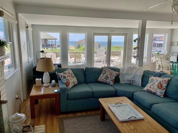 A living room with a teal sofa, patterned cushions, a wooden coffee table, and a view of the beach through large windows. - Sunrise to Sunset - Steps From the Ocean! Hot Tub! (Kitty Hawk)