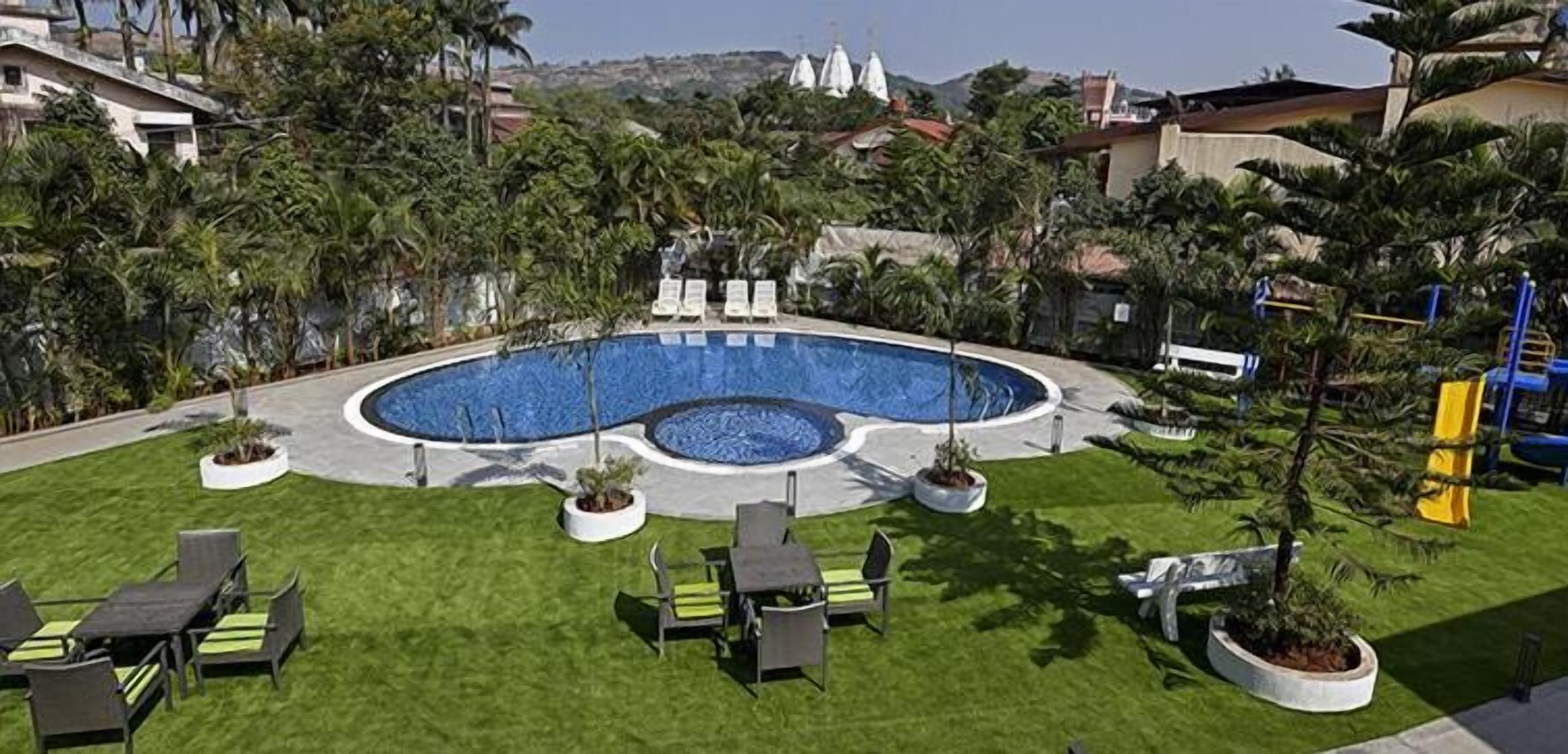 Una piscina con sillas de descanso y jardineras, rodeada de vegetación y edificios al fondo.