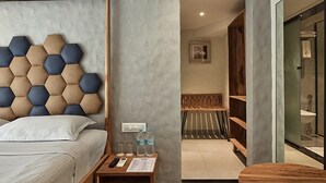 Una habitación de hotel con un cabecero hexagonal, una mesita de noche de madera con un control remoto, un vaso de agua y un banco de madera en el baño.