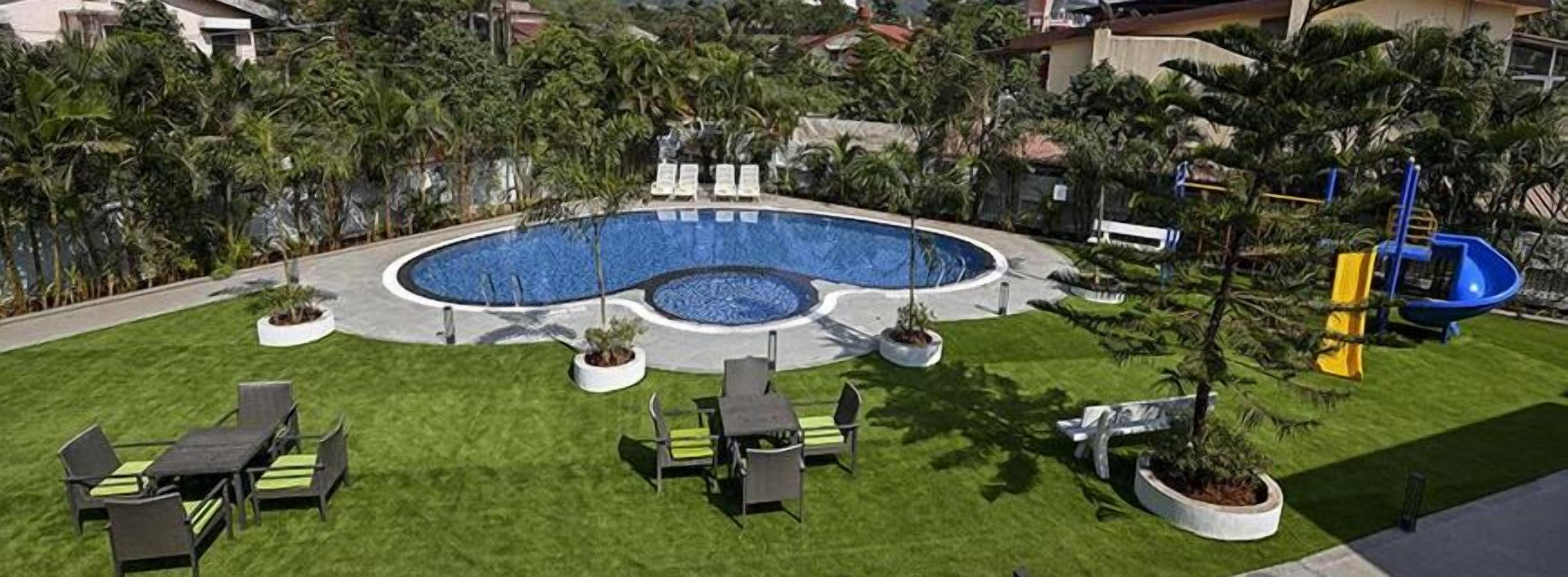 Una piscina con diseño circular, rodeada de césped cuidado, mobiliario exterior y un parque infantil.