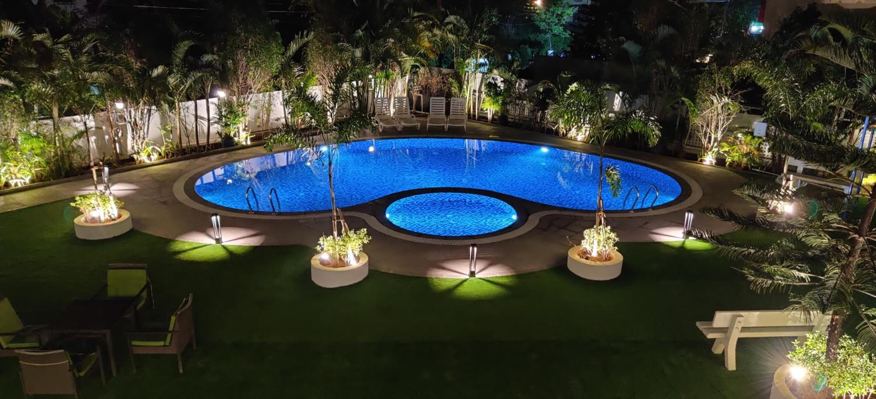 Vista nocturna de una piscina rodeada de vegetación y mobiliario exterior.