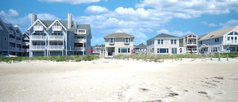 A beachfront property with multiple-story buildings, a sandy beach in the foreground, and a clear sky.