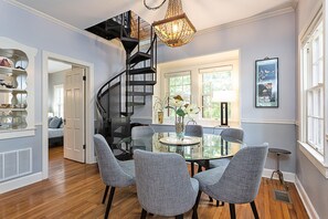 Un coin repas avec une table en verre entourée de quatre chaises grises, un escalier en colimaçon noir et un lustre.
