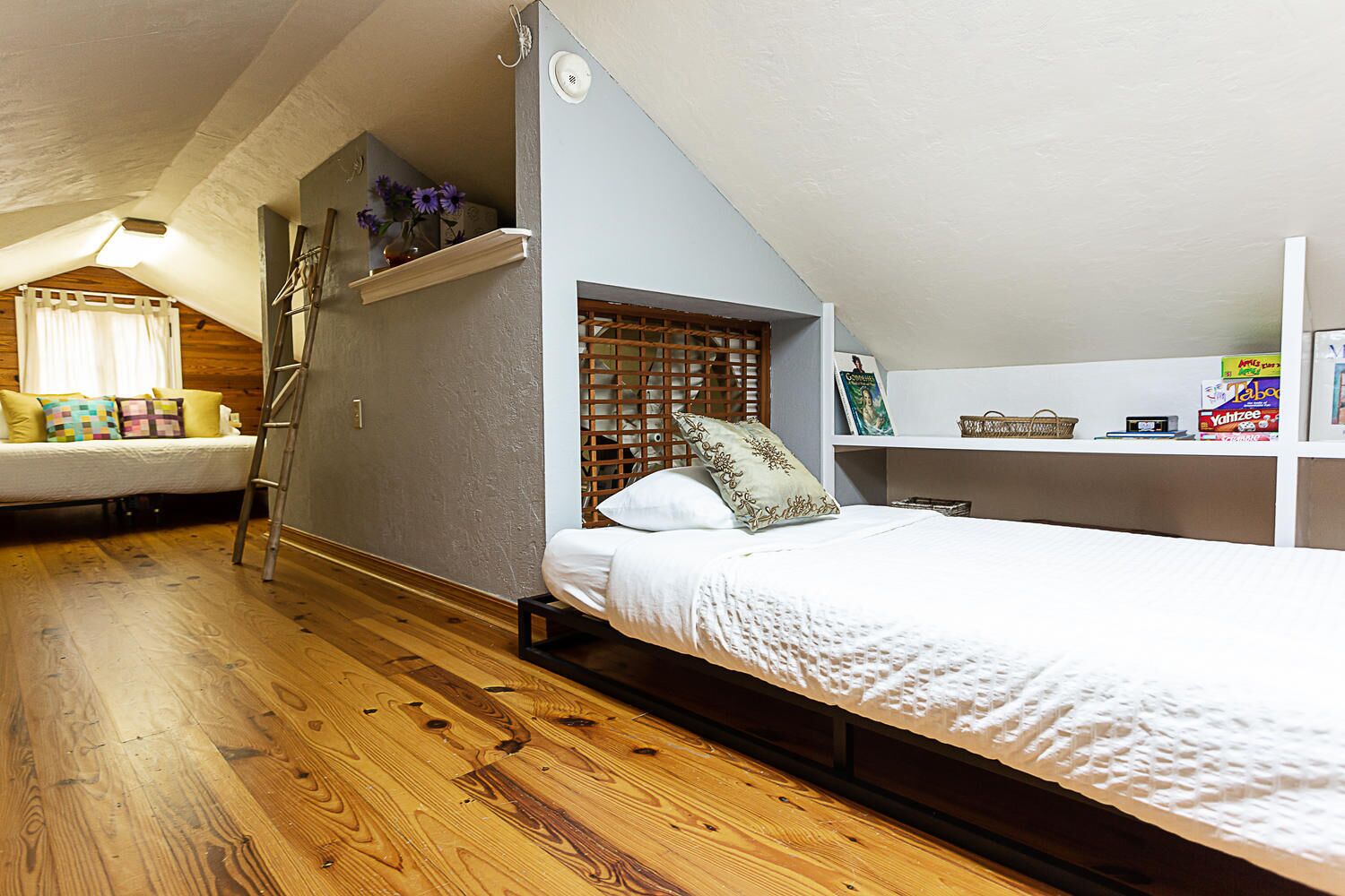 A bedroom with a loft bed, wooden flooring, a small desk with a chair, and a shelf with books and a basket.