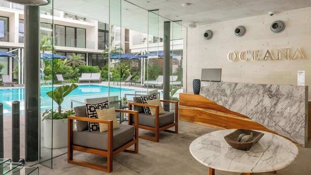 A modern hotel lobby with a pool view, wooden furniture, and a marble reception desk.