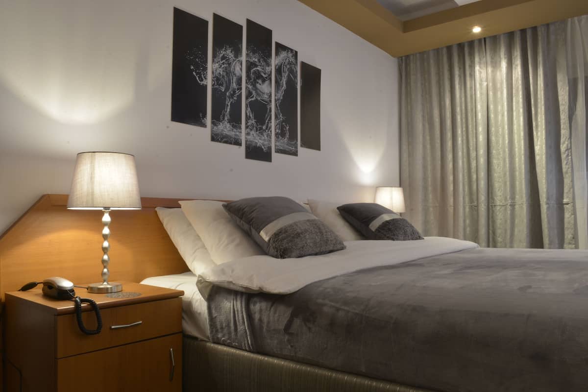Uma cama bem arrumada com edredom cinza e almofadas, uma luminária de cabeceira, mesa de cabeceira de madeira com telefone e uma obra de arte na parede acima da cama.