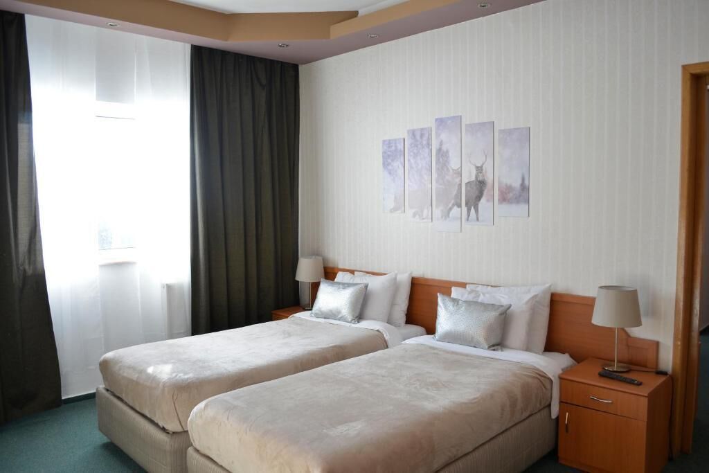 A hotel room with two beds, a wooden headboard, a nightstand with a lamp, and a window with curtains.