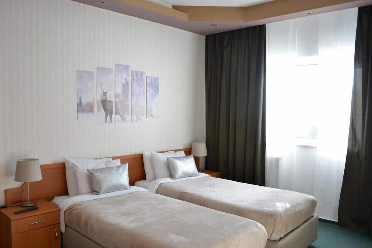 Quarto de hotel com duas camas, uma mesa de cabeceira, uma luminária e uma janela com cortinas.