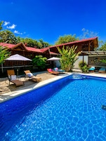 Una piscina con sillas de descanso y sombrillas, rodeada de edificios de madera y vegetación tropical.