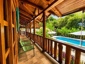 Un balcon en bois donnant sur une piscine, le tout entouré d’une végétation luxuriante.