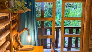 Interior rústico de una cabaña de madera con una mesa, una lámpara de escritorio, un cuaderno y una ventana con vistas a follaje verde.