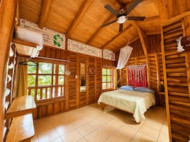 Interior de una cabaña de madera con cama, ventanas, ventilador de techo y suelo de madera.