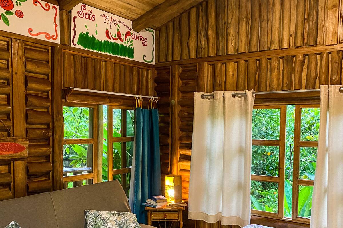 A cozy cabin room with a sofa, a bed with floral bedding, wooden walls, and large windows with curtains.