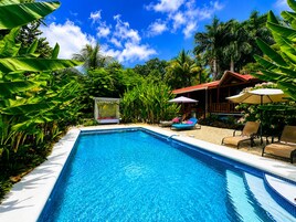 Una piscina con sillas de descanso y una cabaña, rodeada de follaje tropical.