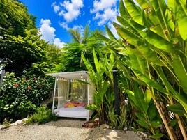 Un jardín con una cama blanca bajo un dosel, rodeado de exuberante follaje y plantas de banano.