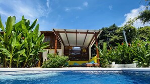 Un área de piscina con zona de estar cubierta, abundante vegetación y cielo azul despejado.