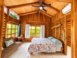 A wooden cabin room with a bed, a sofa, a ceiling fan, and a window with curtains. - Cabañas Lamaite (Sámara)