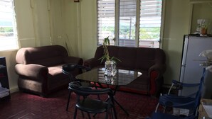 A living room with a sofa, a glass coffee table, and a chair. There is a window with blinds, a refrigerator, and a plant on the table. - Matus Guest House (San Ignacio)