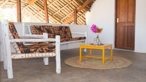 Una habitación con techo de paja, puerta de madera, sofá blanco con cojines estampados y una pequeña mesa amarilla con un florero.