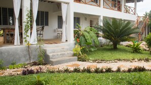Un edificio de dos pisos con un porche cubierto, mobiliario exterior y un jardín bien cuidado con plantas tropicales.